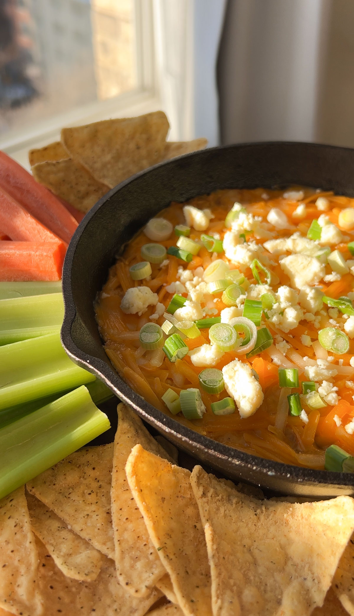 A skillet of vegan buffalo dip garnished with green onions on a platter with veggie sticks.