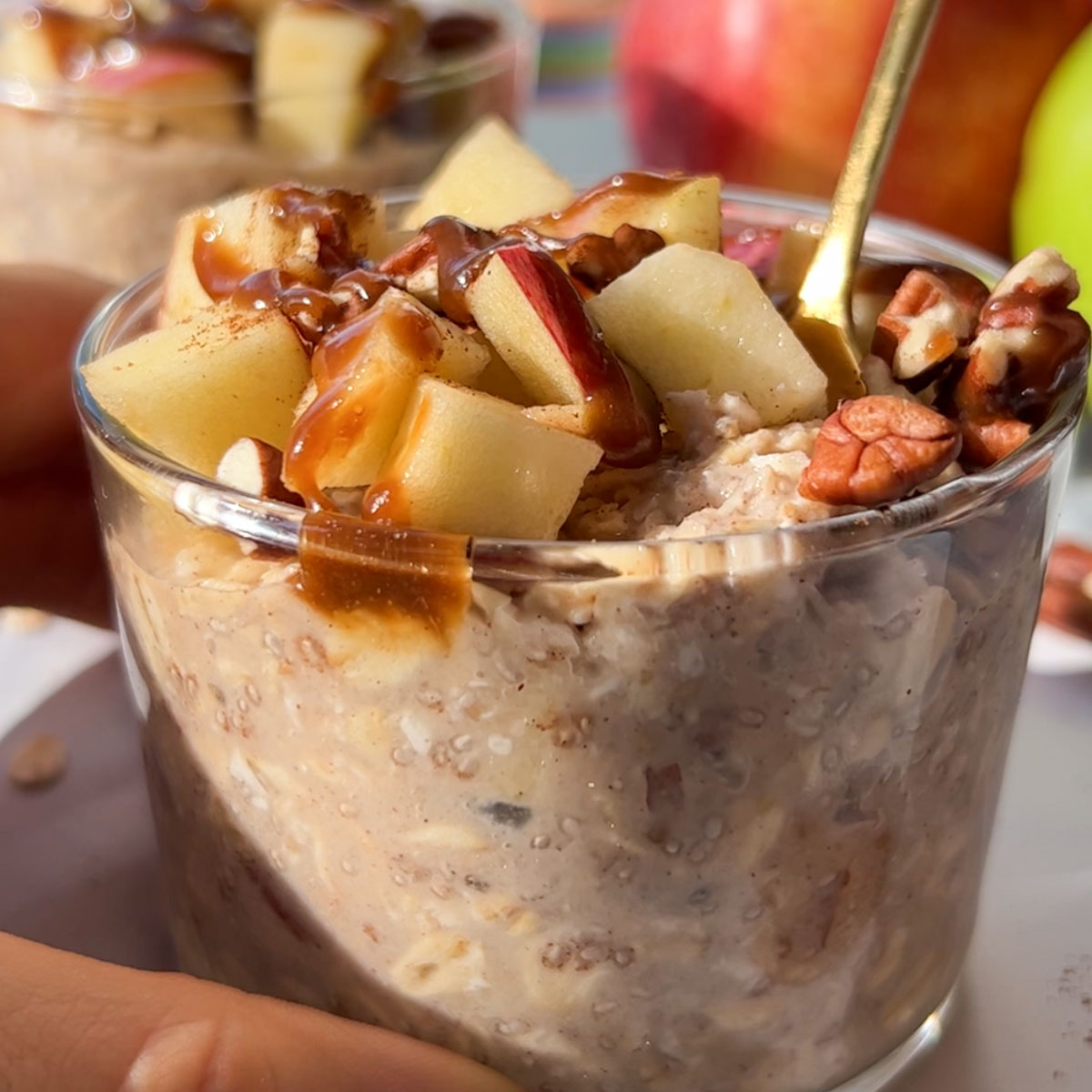 A spoon dipping into apple overnight oats in a jar topped with pecans, apple pieces, and caramel sauce.