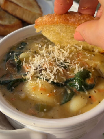 A bowl of vegan gnocchi soup with a hand dipping a piece of bread into the bowl.