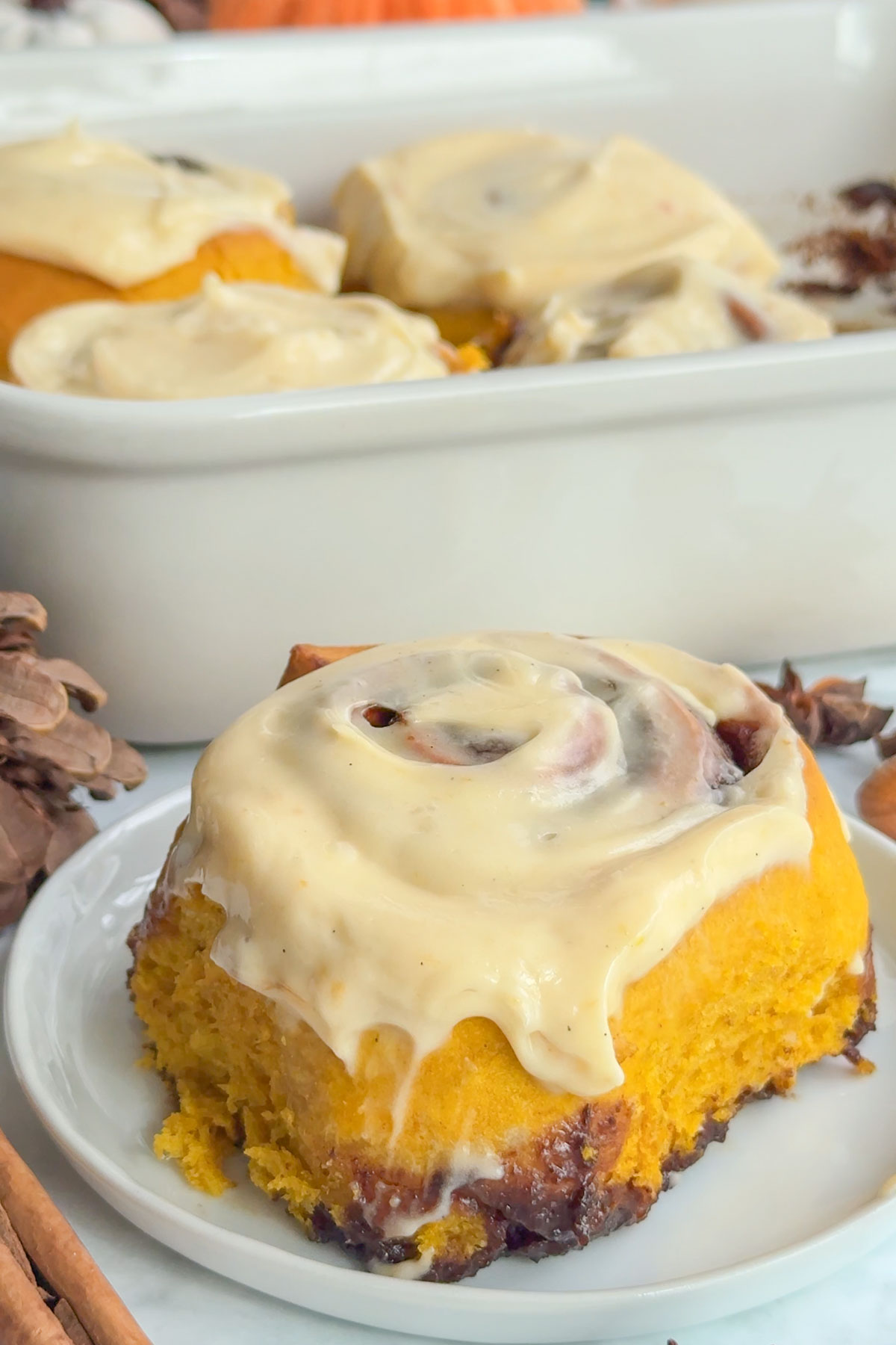 Pumpkin cinnamon roll on a plate with more in a baking dish behind it. 