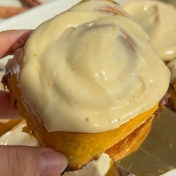 A hand holding up a pumpkin cinnamon roll with icing.