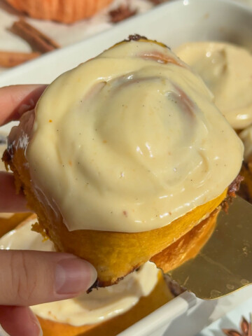 A hand holding up a pumpkin cinnamon roll with icing.
